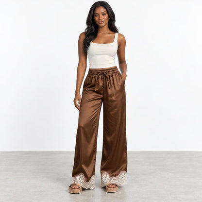 Woman in a white tank top and brown satin pants on a plain background.