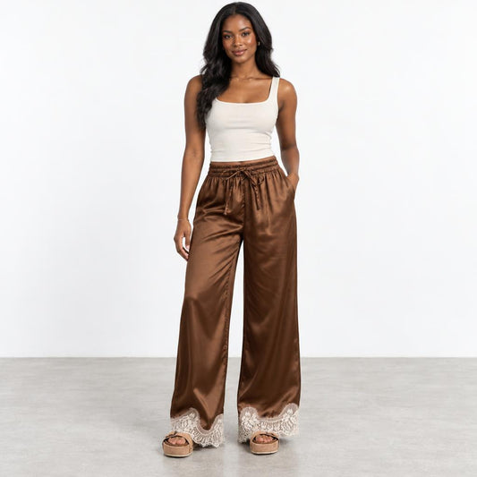 Woman in a white tank top and brown satin pants on a plain background.