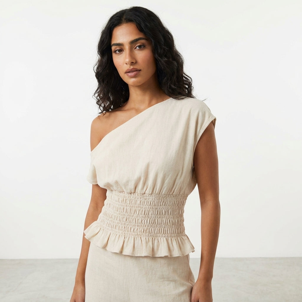 Woman in a beige, one-shoulder, ruffled top against a plain backdrop.