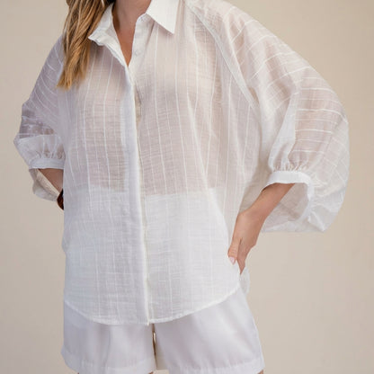 Woman in a sheer white blouse and shorts, hand on hip.