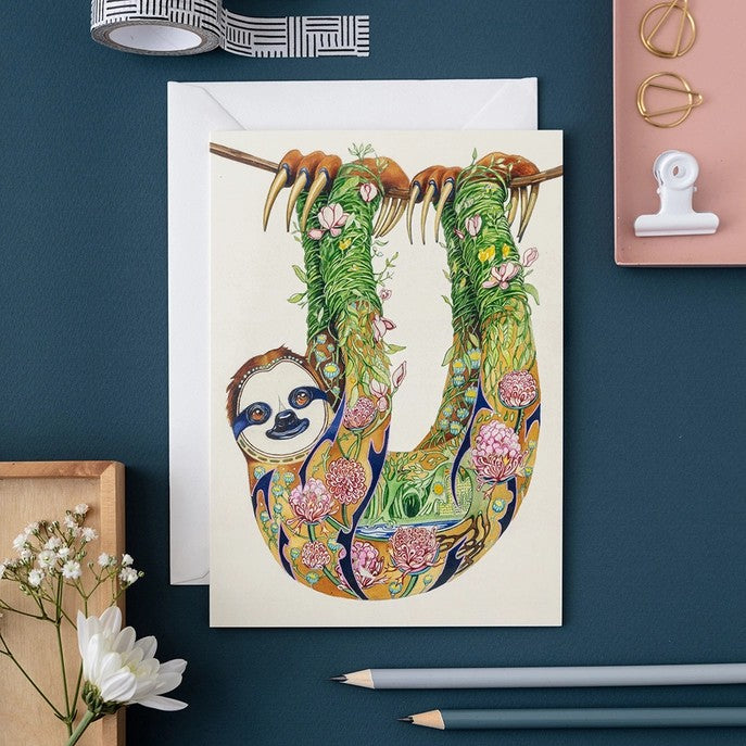Illustrated sloth card with floral design on a desk.