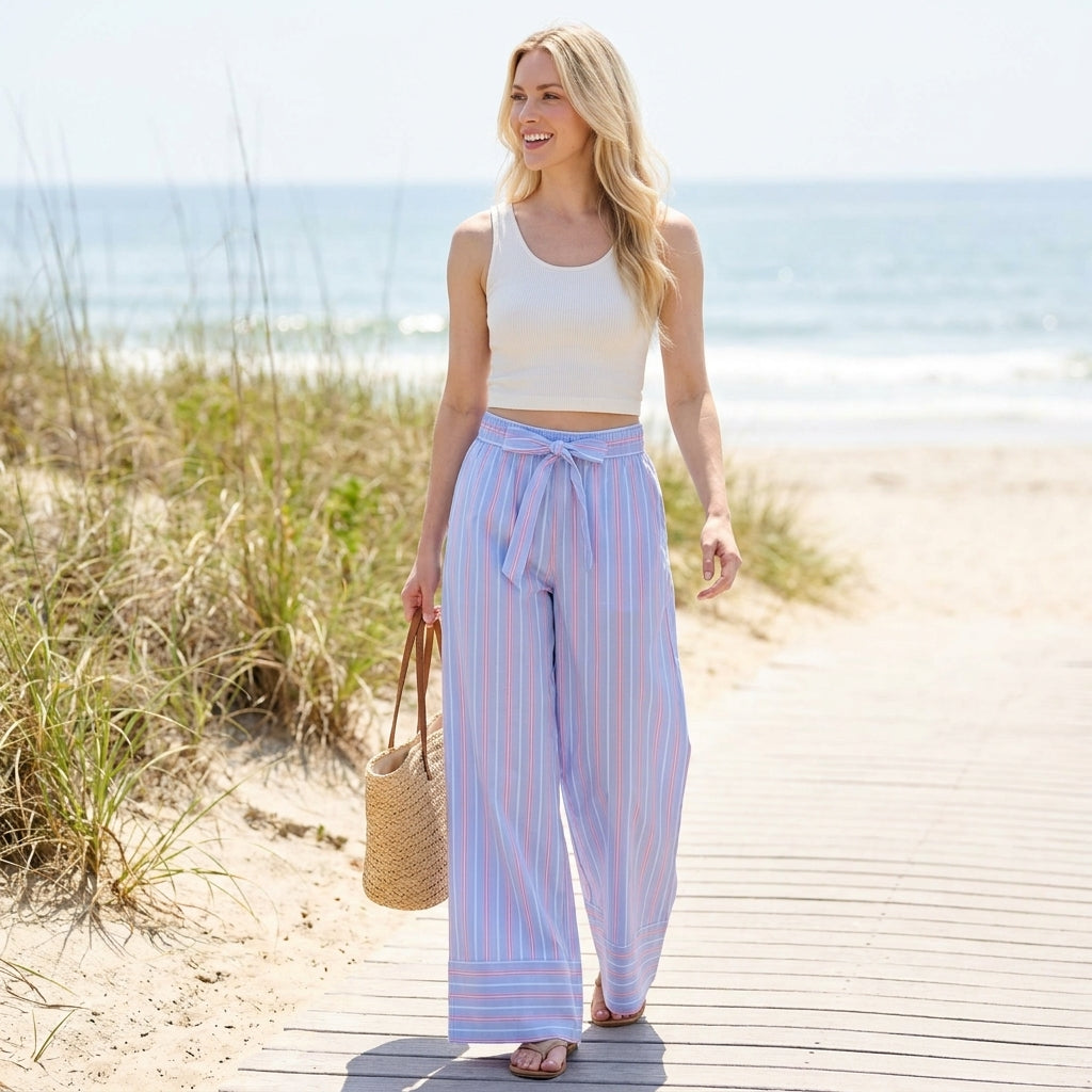 Woman in striped pants walking on a beach boardwalk, holding a straw bag.
