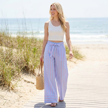 Woman in striped pants walking on a beach boardwalk, holding a straw bag.