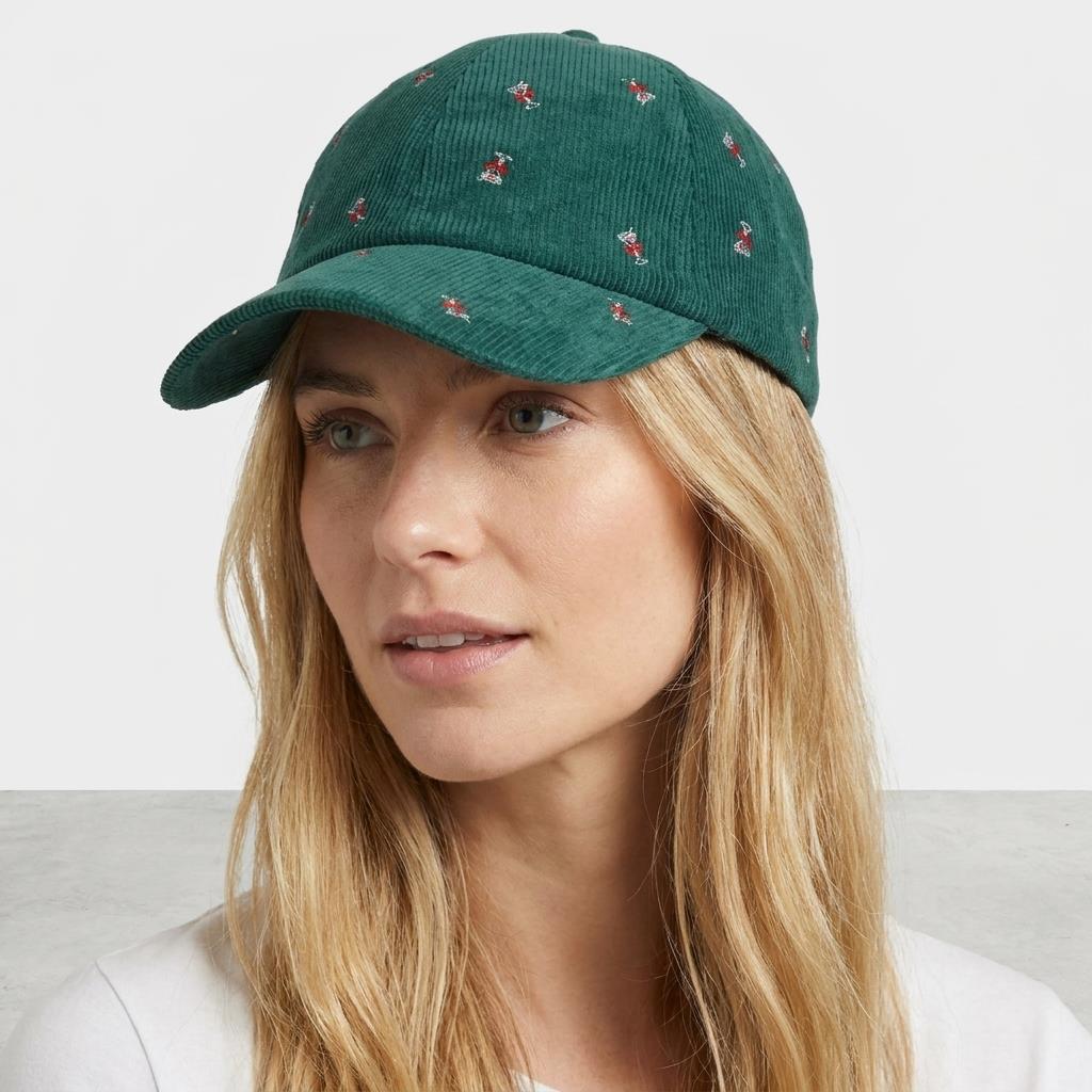 A woman models a green embroidered corduroy cap against a plain studio backdrop.