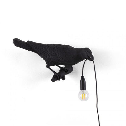 Black bird sculpture holding a lightbulb by a wire in its beak.