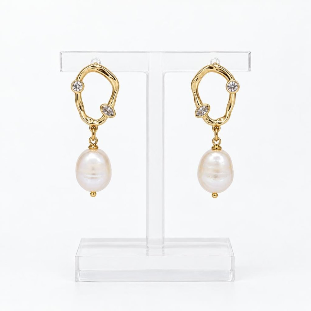 Gold-tone drop earrings with sparkling stones and pearl pendants displayed on a clear stand.