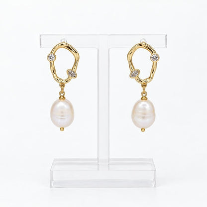 Gold-tone drop earrings with sparkling stones and pearl pendants displayed on a clear stand.