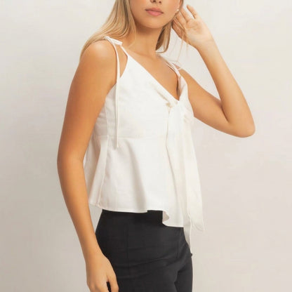 Woman in white sleeveless top and black pants, posing with hand near ear.