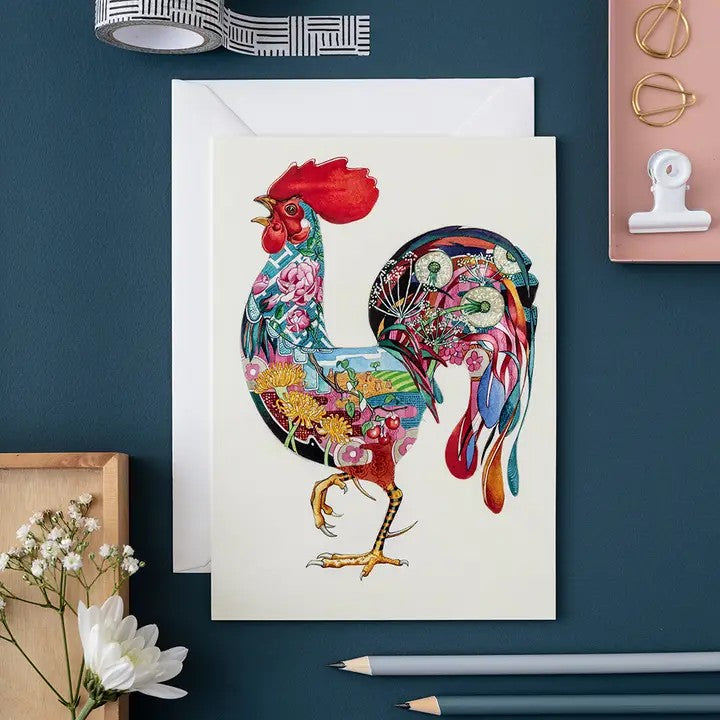 Colorful rooster card with floral details on a dark desk.