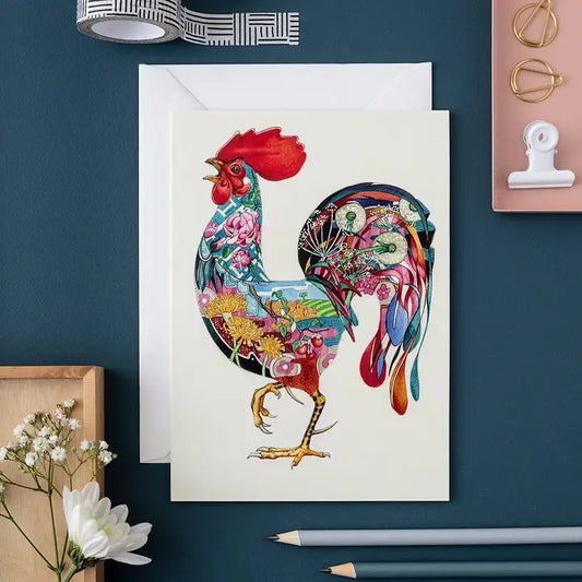 Colorful rooster card with floral details on a dark desk.