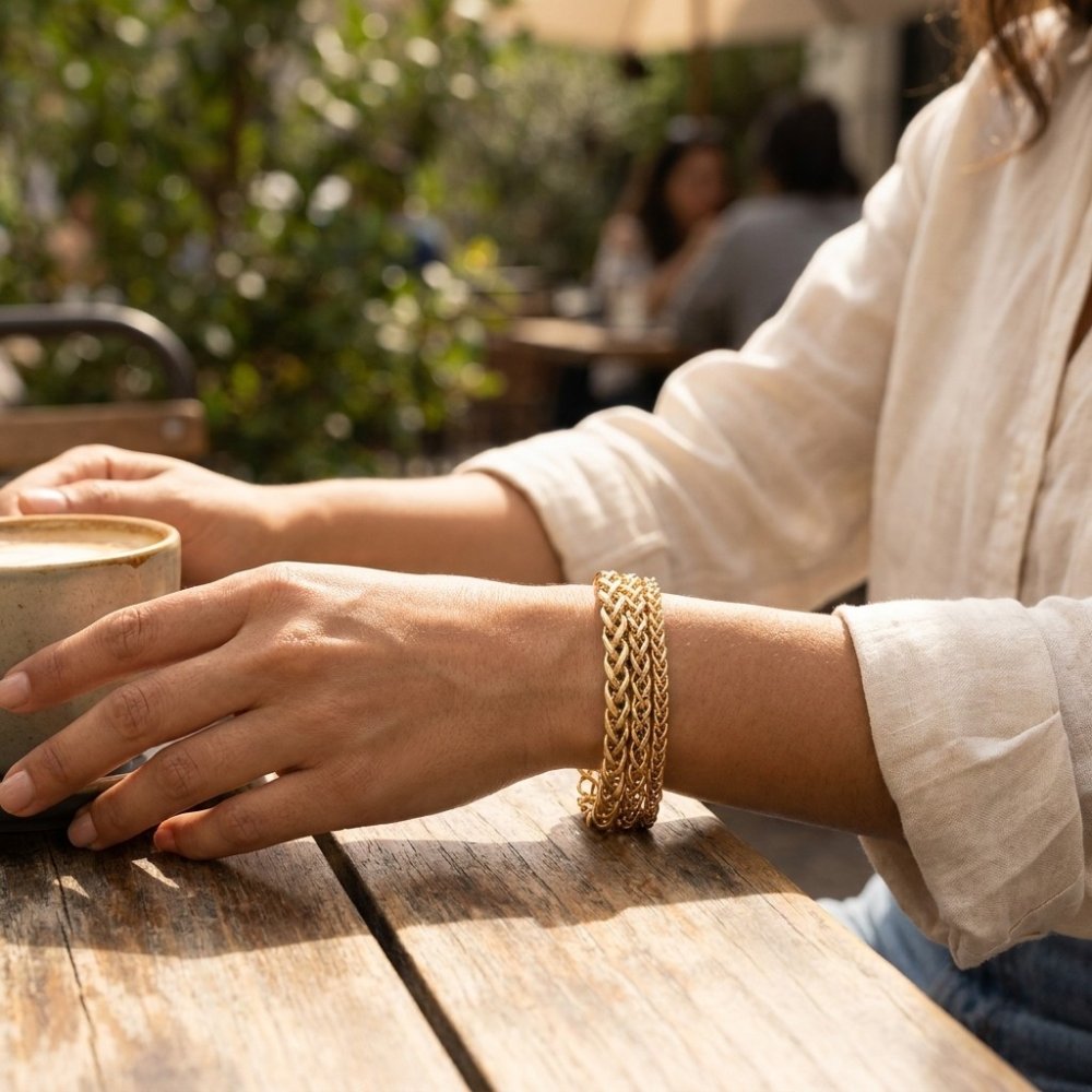 A person wearing a gold bracelet holds a coffee cup at a sunlit outdoor café.