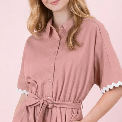 Woman in a pink button-up dress with a waist tie, standing against a soft pink background.