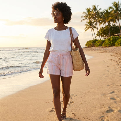 Woman in white tee walks on beach at sunset, carrying a straw bag.