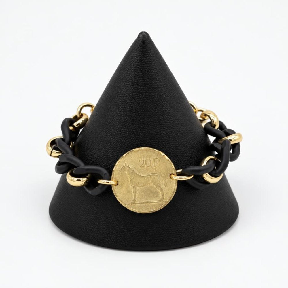 A black and gold chain bracelet with a coin charm displayed on a black stand.