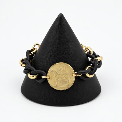A black and gold chain bracelet with a coin charm displayed on a black stand.