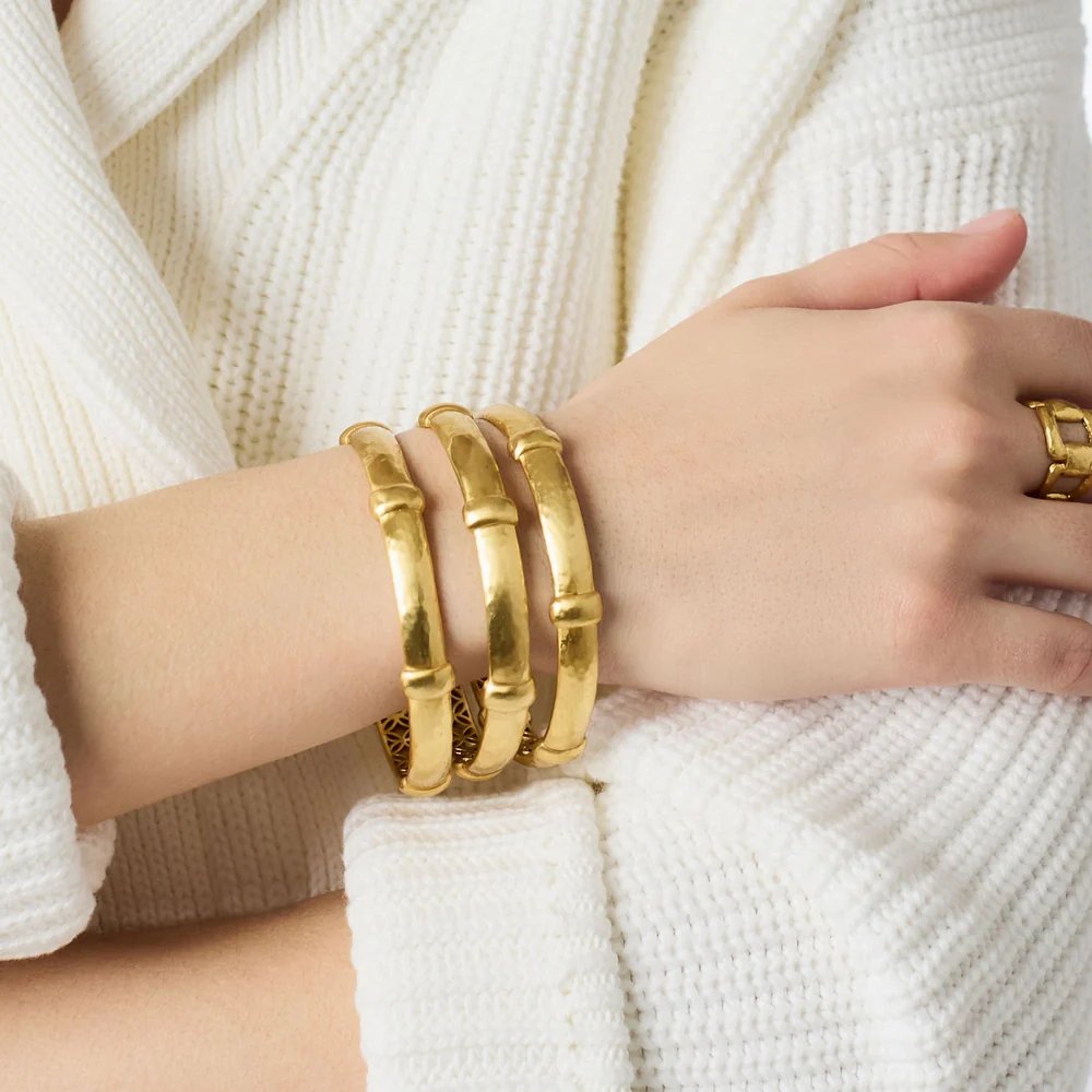 A hand wearing three gold bamboo-style bangles and a matching ring over a white knit sweater.