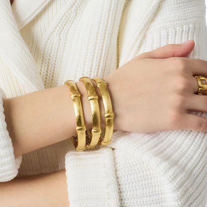 A hand wearing three gold bamboo-style bangles and a matching ring over a white knit sweater.