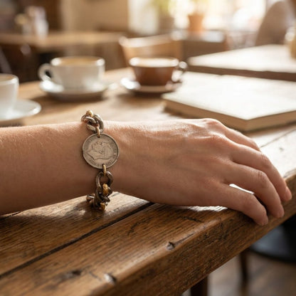 A hand wearing a coin charm bracelet rests on a wooden café table beside cups and a book.