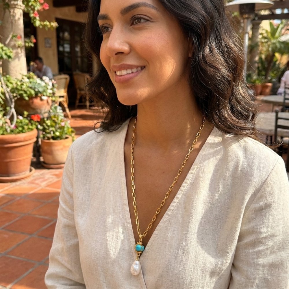 A smiling woman in a white blouse and pendant necklace sits in a sunny garden café.