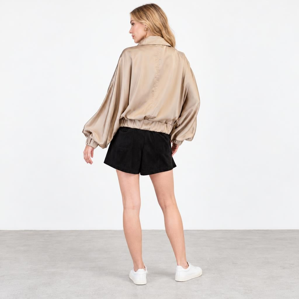 A woman models a beige satin jacket, black shorts, and white sneakers against a studio backdrop.
