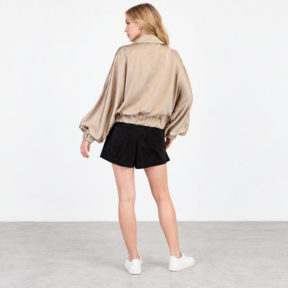 A woman models a beige satin jacket, black shorts, and white sneakers against a studio backdrop.