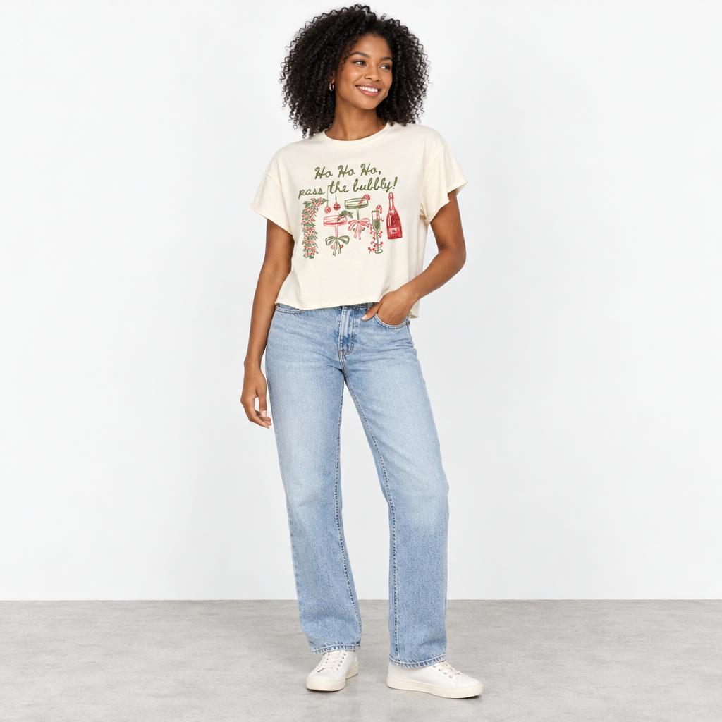 A smiling woman models a festive graphic T-shirt with light-wash jeans and white sneakers.