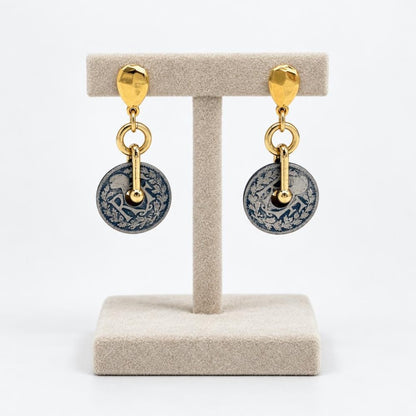 Gold-tone drop earrings with ornate blue and silver circular pendants on a display stand.