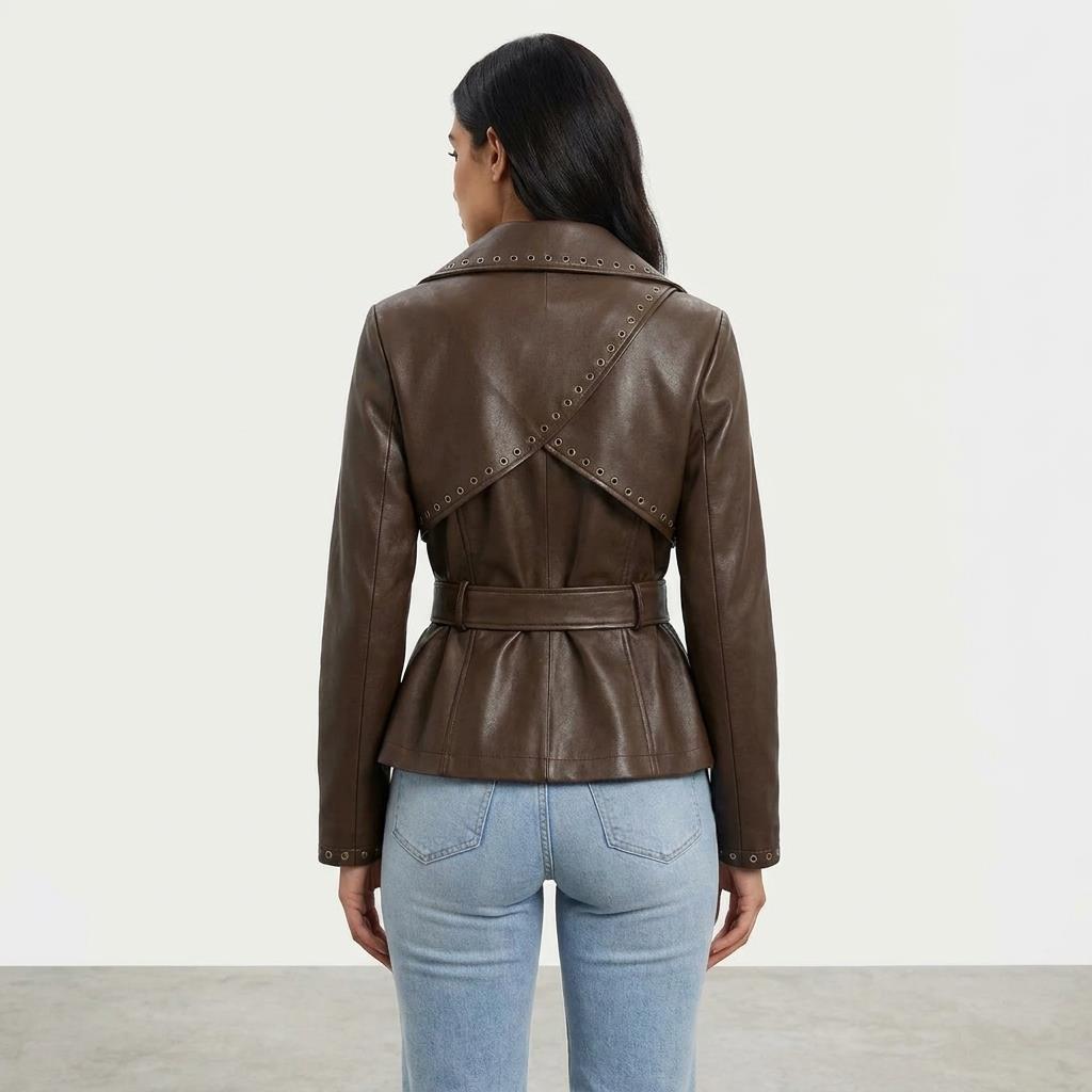 A woman models a belted brown leather jacket with decorative eyelet detailing and light blue jeans.