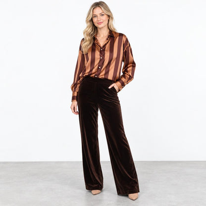 A woman models a striped satin blouse and dark brown velvet wide-leg pants in a studio.