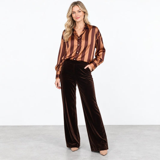 A woman models a striped satin blouse and dark brown velvet wide-leg pants in a studio.