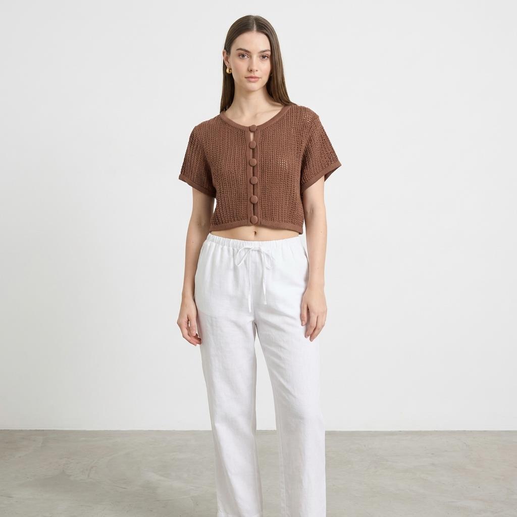 Woman in brown knit top and white pants standing against a plain background.