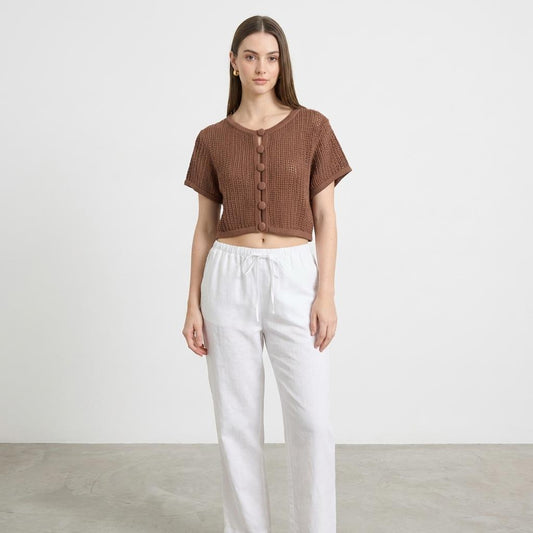 Woman in brown knit top and white pants standing against a plain background.