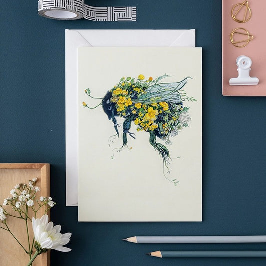 Bee art card with flowers, pencils, and floral decor on a blue surface.