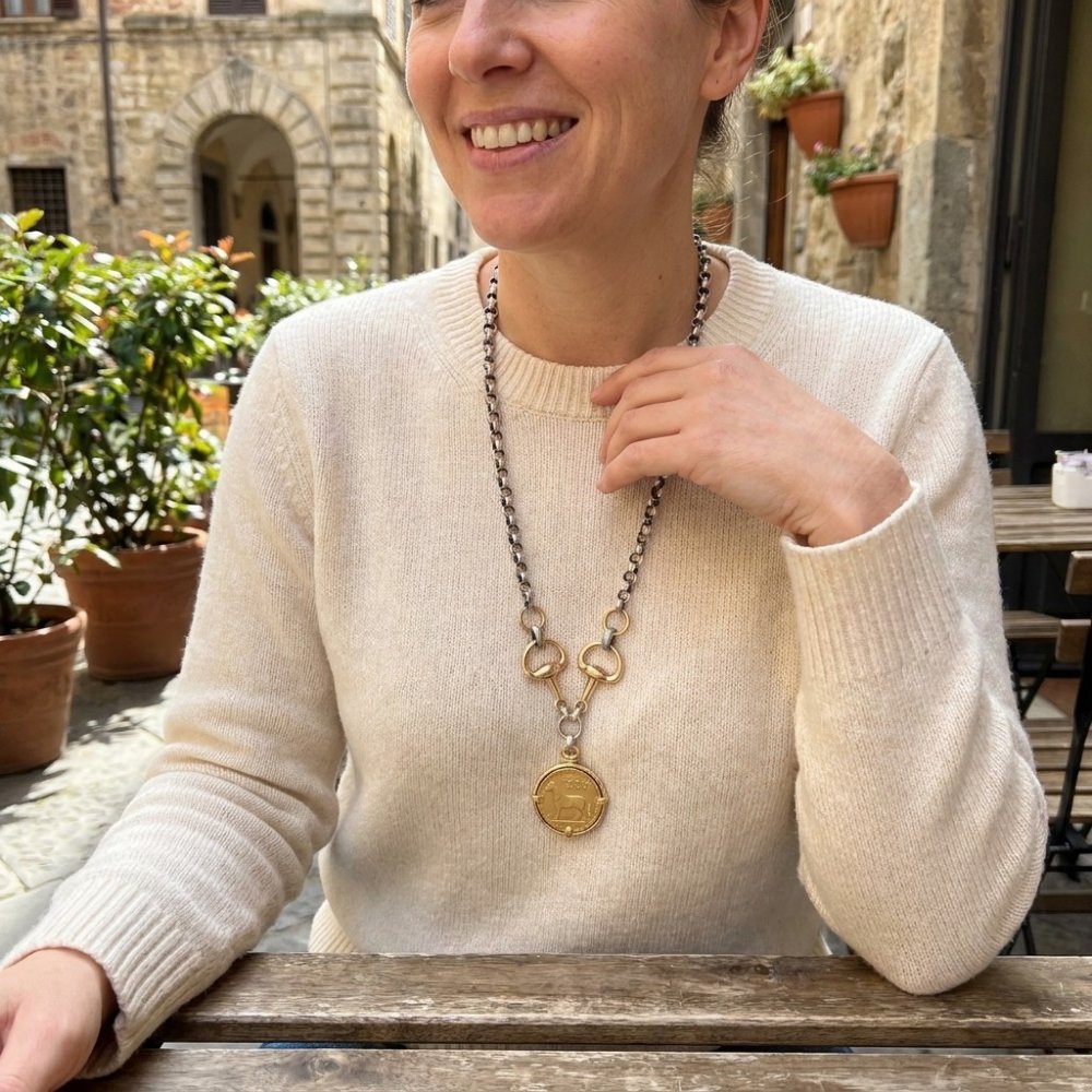 A smiling woman in a cream sweater showcases a gold pendant necklace at an outdoor café.