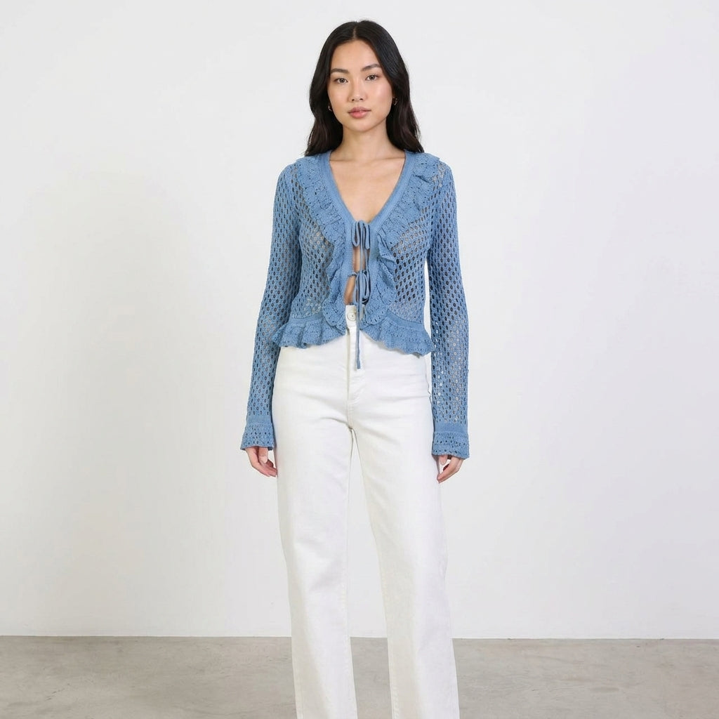 Woman in a blue crochet top and white pants stands against a plain background.