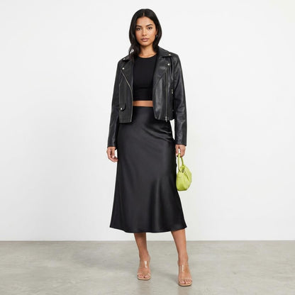 Woman in black outfit with leather jacket and green bag, standing on a gray floor.