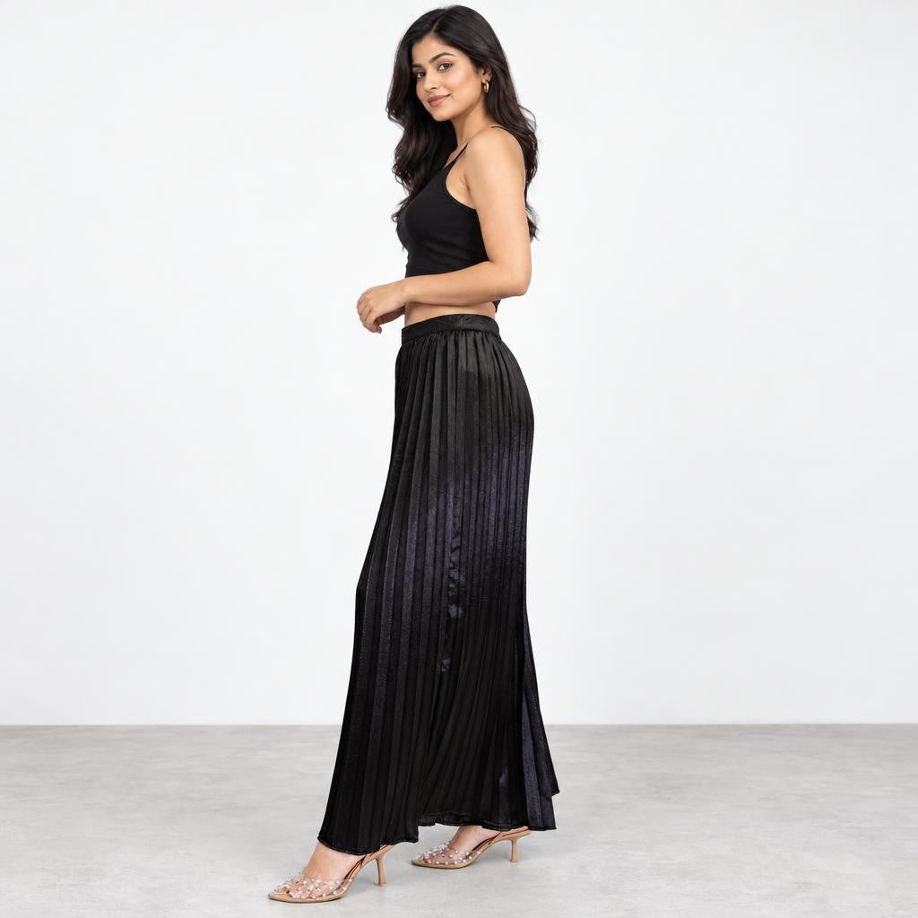 A woman models a black crop top and pleated maxi skirt against a plain studio backdrop.