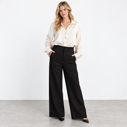 A woman models a white blouse and black wide-leg trousers in a minimalist studio.