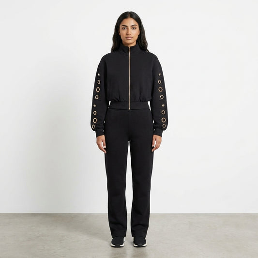 Woman in a black outfit with grommeted sleeves standing on a plain background.