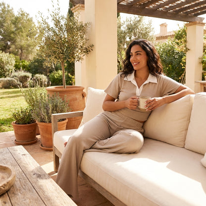 Woman relaxing with coffee on a patio sofa, surrounded by lush greenery.