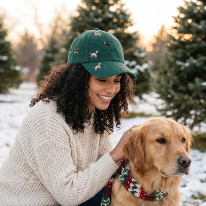 A woman in a green cap pets a golden retriever wearing a festive scarf in a snowy park.