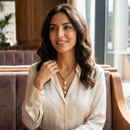 A smiling woman in a white blouse wears a gold necklace with pearl accents in a sunlit café.