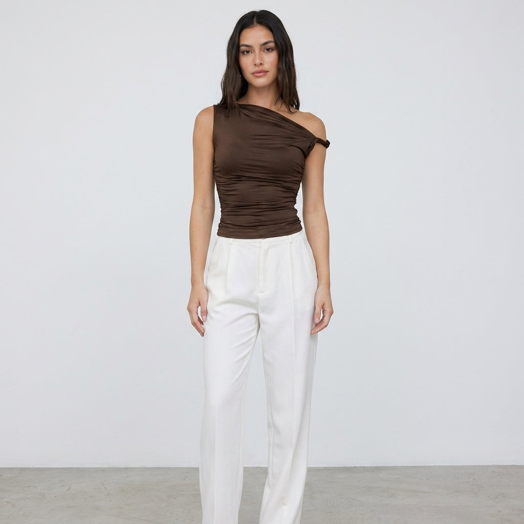 Woman in brown one-shoulder top and white pants against plain background.