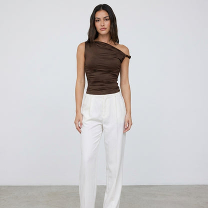 Woman in brown one-shoulder top and white pants against plain background.