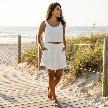 Woman in white outfit walks on the beach, smiling under the sun.