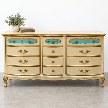 An ornate cream-and-gold dresser with turquoise accents, topped with books and a leafy plant.