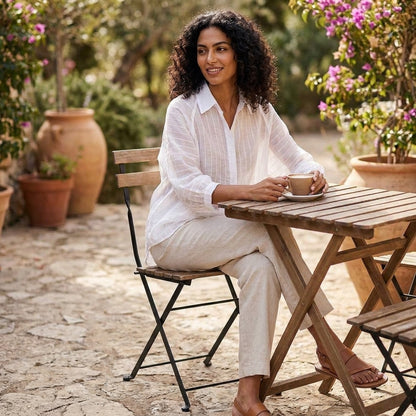 Woman enjoys coffee at a wooden table surrounded by greenery and flowers.
