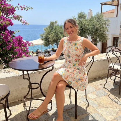 Woman smiling, enjoying a drink by the sea with vibrant flowers and blue skies.