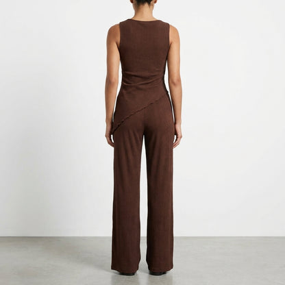 Woman in brown sleeveless jumpsuit, facing away, with simple background.