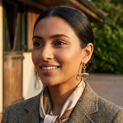 A smiling woman in a tweed coat and silk scarf stands in warm evening sunlight.